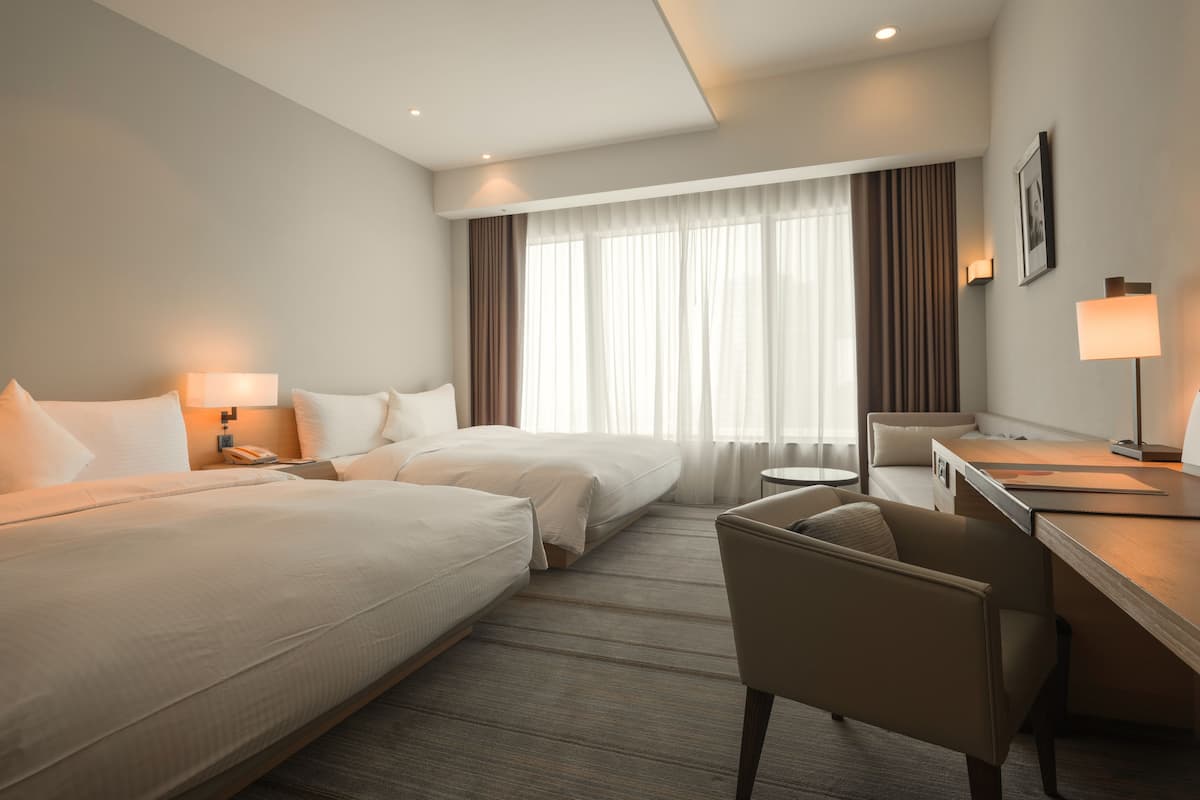 Une chambre d’hôtel avec deux lits, un bureau, une chaise, une petite table ronde et une fenêtre avec des rideaux.