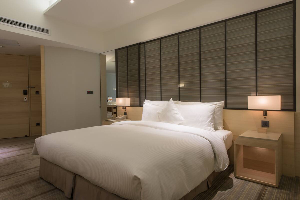 Une chambre d’hôtel moderne avec un grand lit, une tête de lit ornée de lignes horizontales, des lampes de chevet et un plancher en bois.