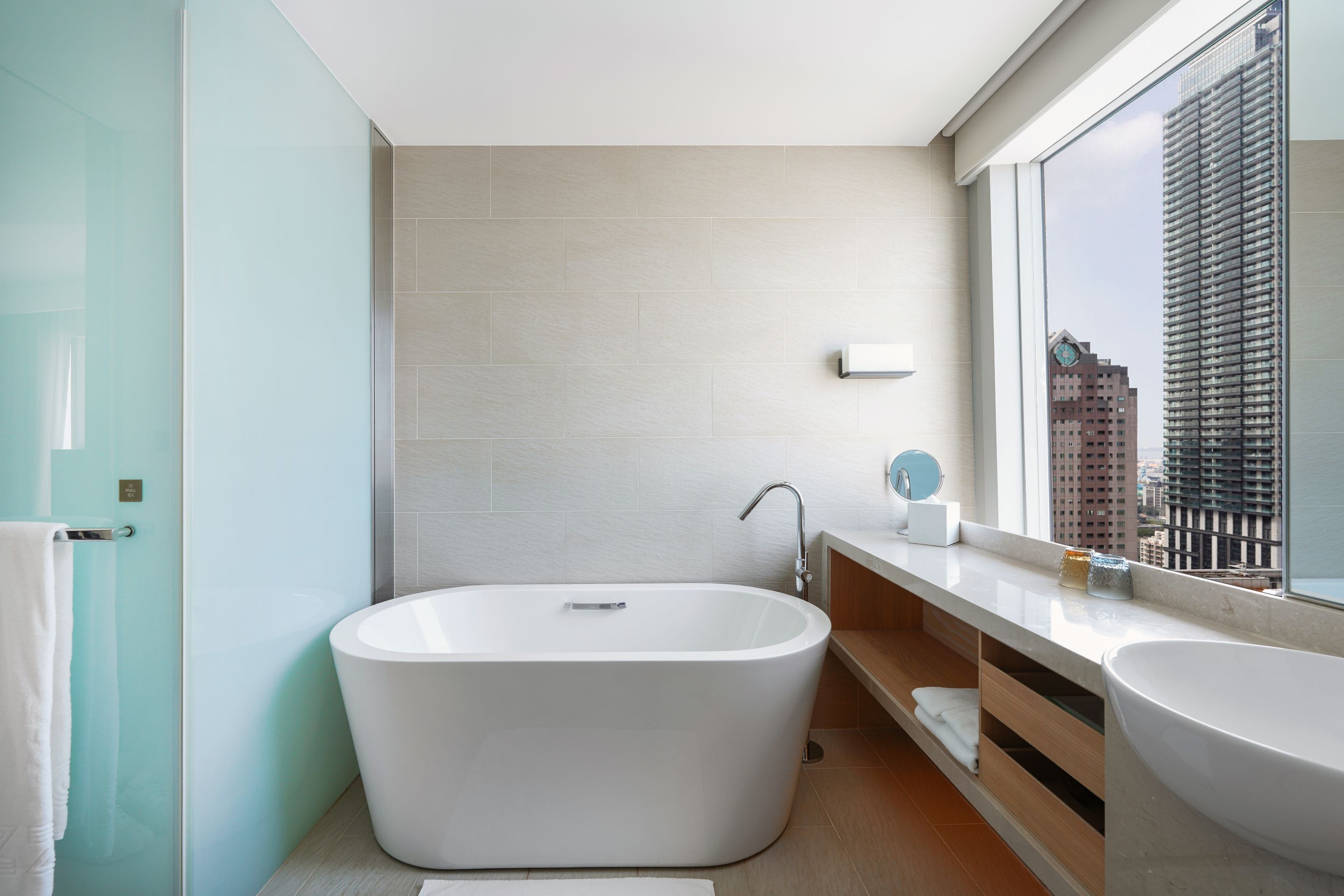 Un baño moderno con una bañera independiente, un tocador con espejo y un amplio ventanal con vistas a la ciudad.