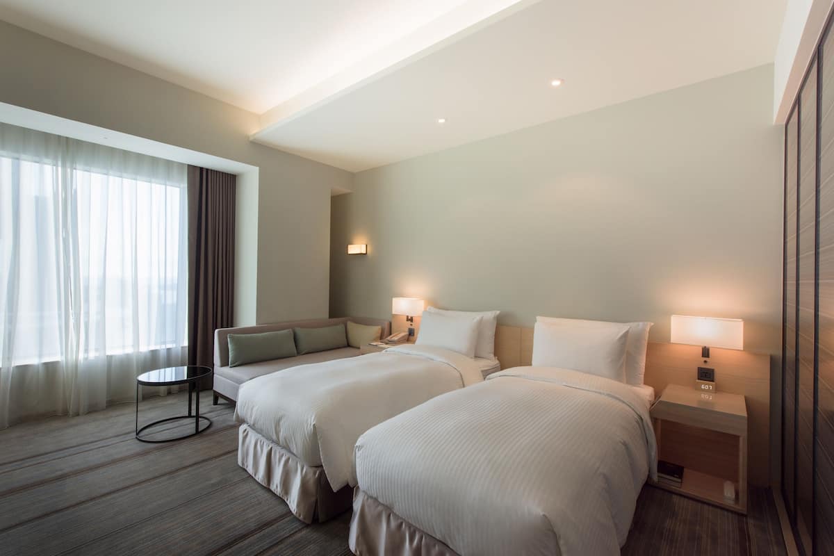 Une chambre d’hôtel avec deux lits, une petite table, une table de chevet avec une lampe et une fenêtre avec des rideaux.
