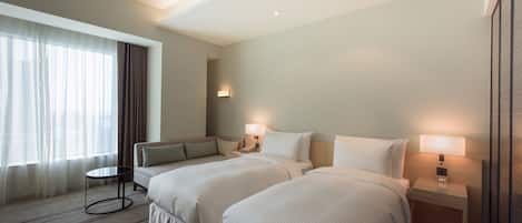 Habitación de hotel con dos camas, una mesita pequeña, una mesita de noche con lámpara y una ventana con cortinas.
