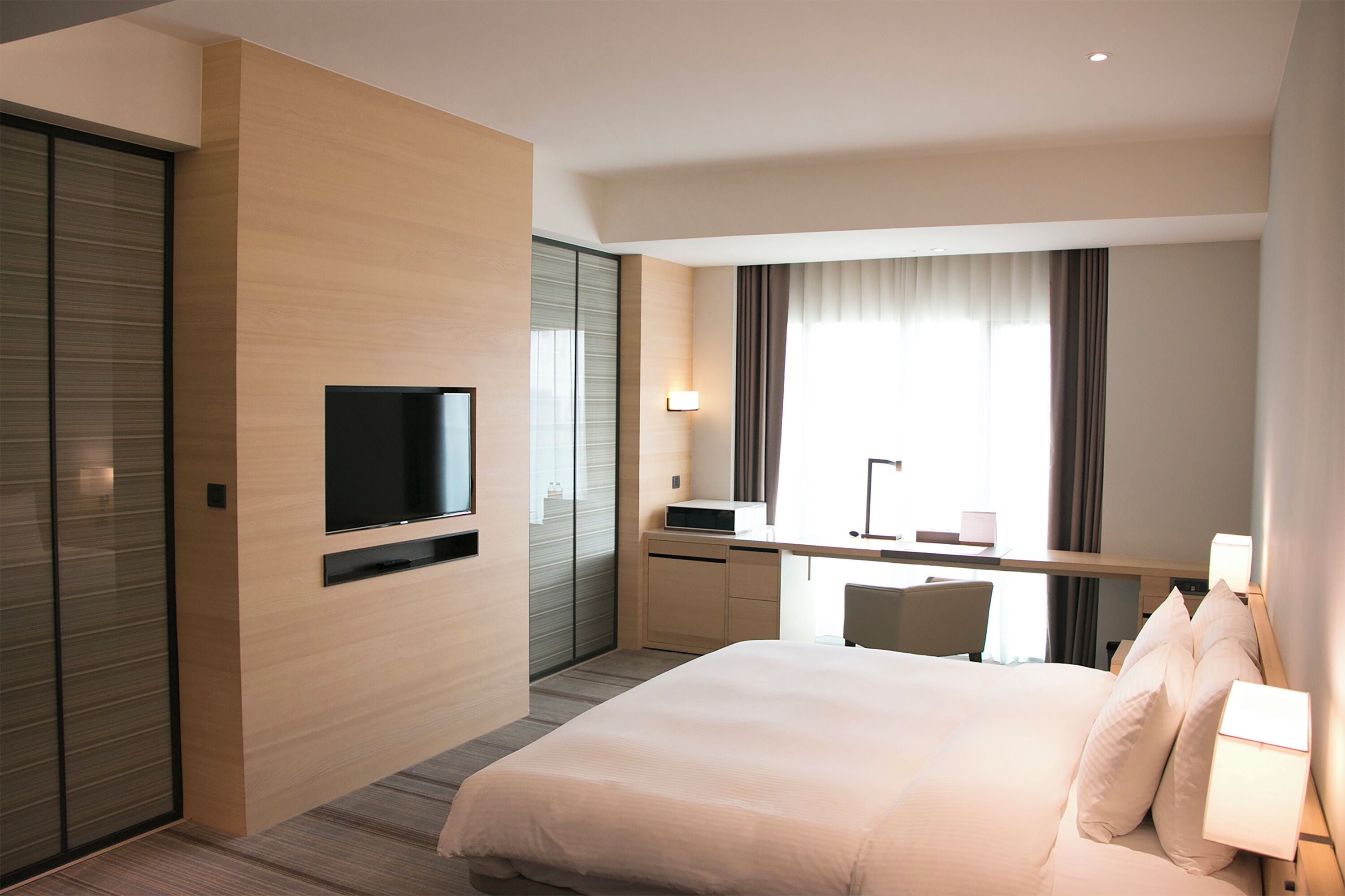 A modern hotel room with a large bed, a flat-screen TV mounted on the wall, a desk with a chair, and a large window with curtains.