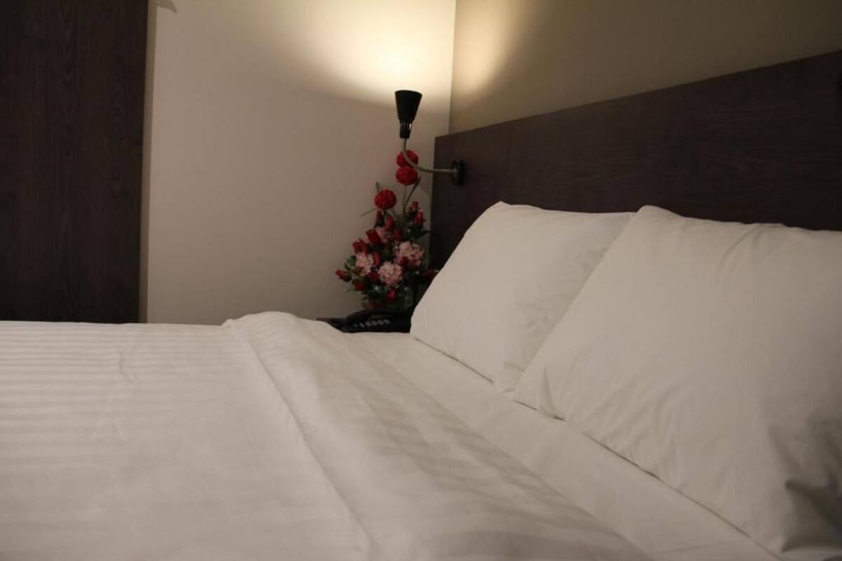 Una cama bien hecha con sábanas blancas, cabecera de madera oscura y lámpara de pared con diseño floral.