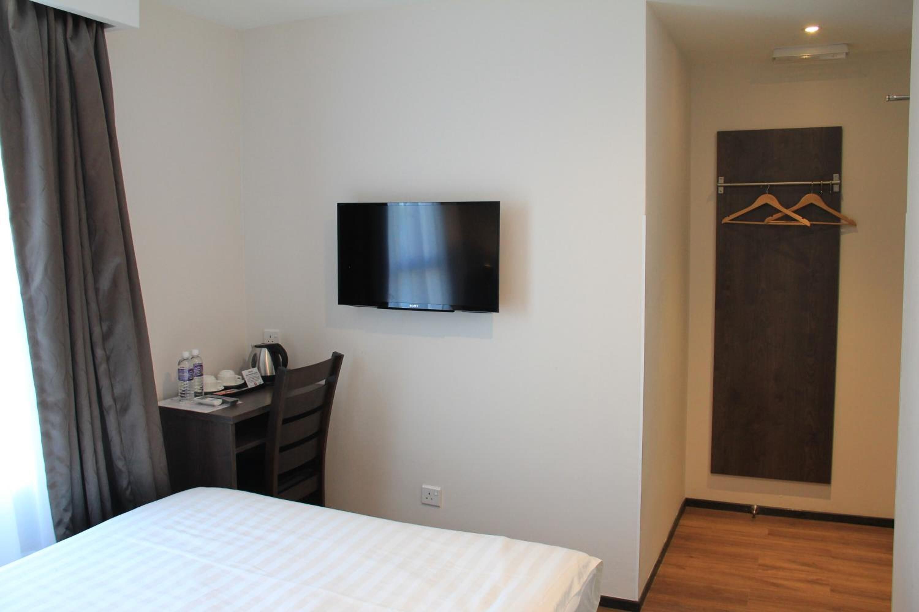 A hotel room with a bed, a desk with a chair, a television mounted on the wall, and a coat rack with hangers.