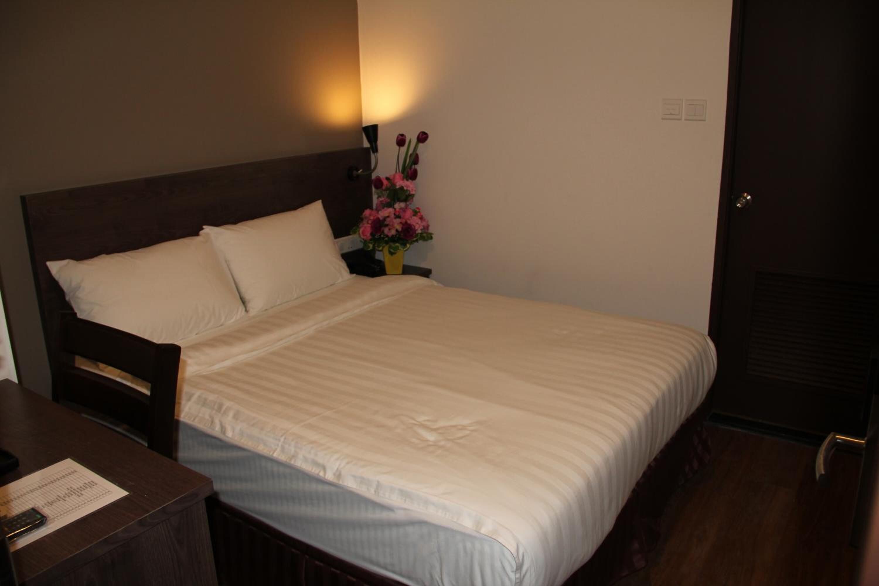 A neatly made bed with white linens, a wooden headboard, a nightstand with a lamp and a vase of flowers, and a door with a handle.