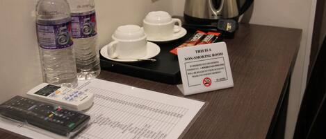 Habitación de hotel con una mesa dispuesta con dos botellas de agua, dos tazas blancas, una tetera, un control remoto y un cartel de no fumar.
