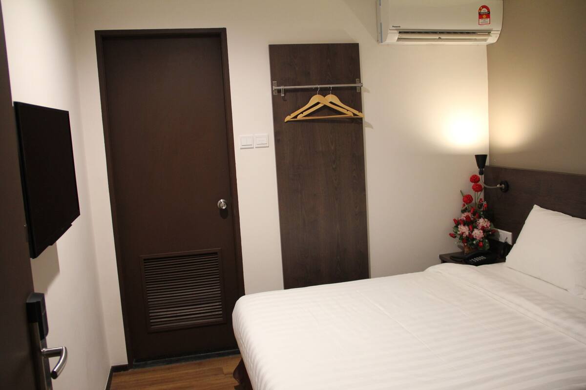 Una habitación de hotel con una cama, una puerta de madera, aire acondicionado instalado en la pared, una mesita de noche con lámpara y un pequeño arreglo floral.