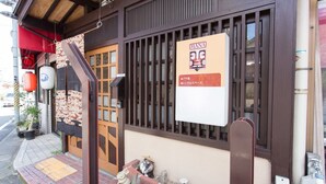 Edificio japonés tradicional con puerta de madera, un letrero que dice 'MANA' y 'プチ宿' indicando una posada pequeña, y un jardín con piedras decorativas y un farol.