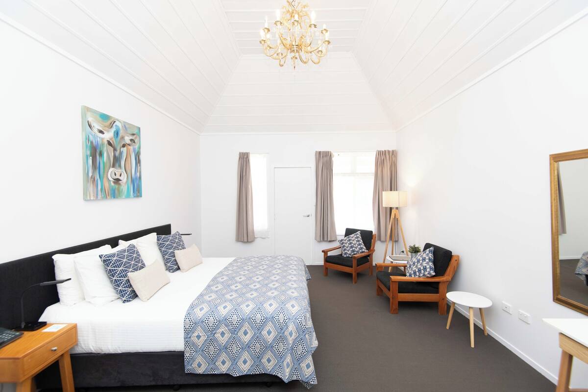 Ein Hotelzimmer mit einem Bett, einem Schreibtisch, einem Sessel, einem kleinen Tisch, einer Lampe, einem Gemälde und einer Kandelaber.