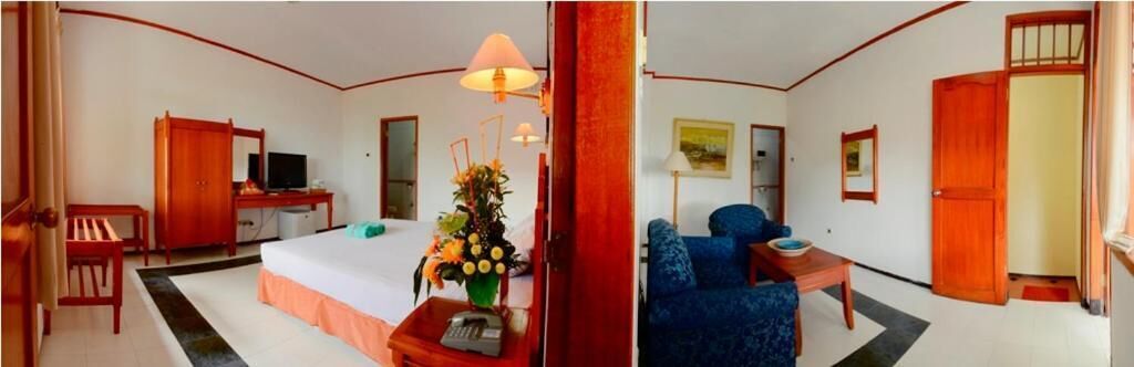A hotel room with a bed, desk, chair, TV, and a small table with a lamp and flowers.