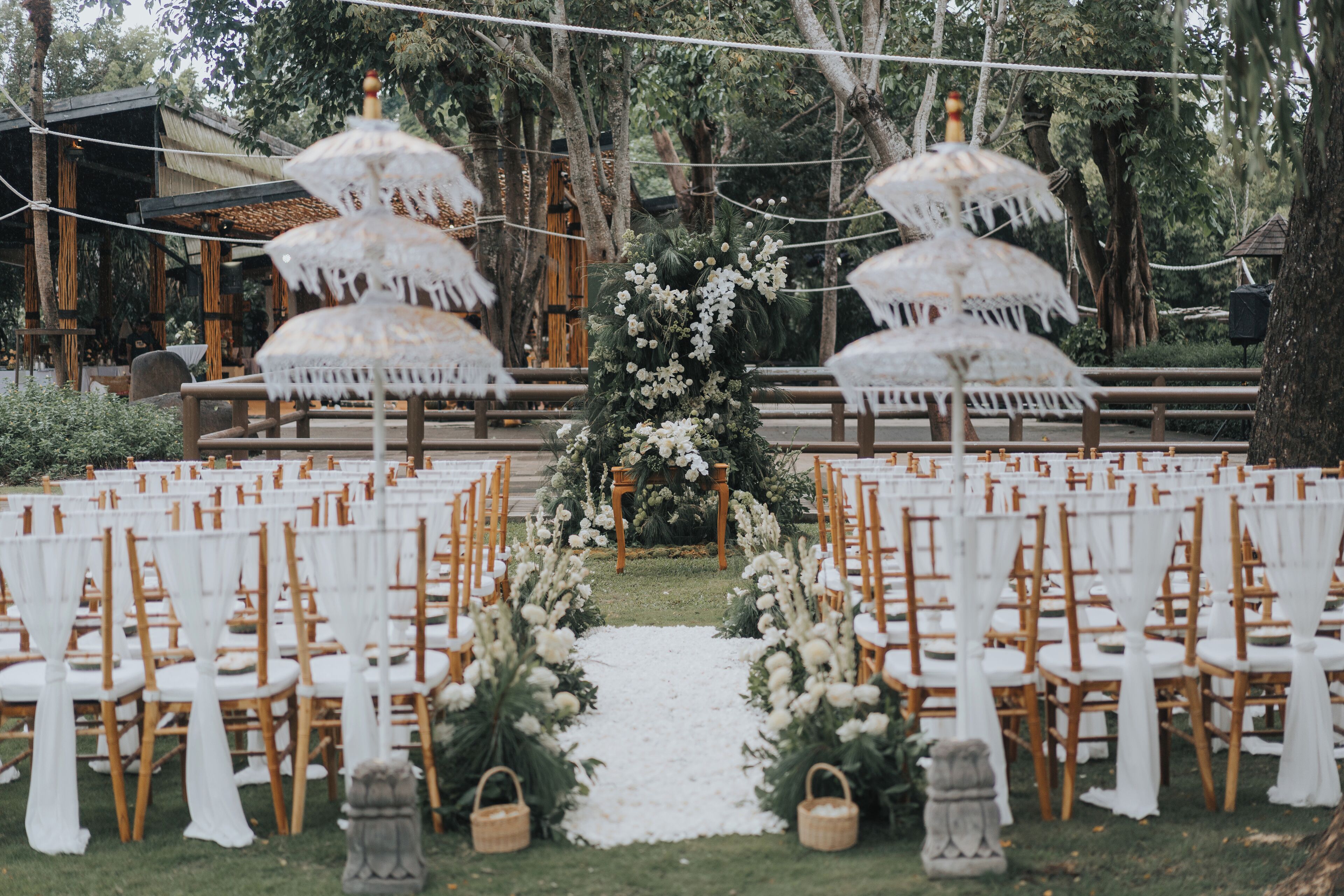 An outdoor wedding venue with rows of wooden chairs and white tablecloths, a floral archway, and decorative umbrellas.
