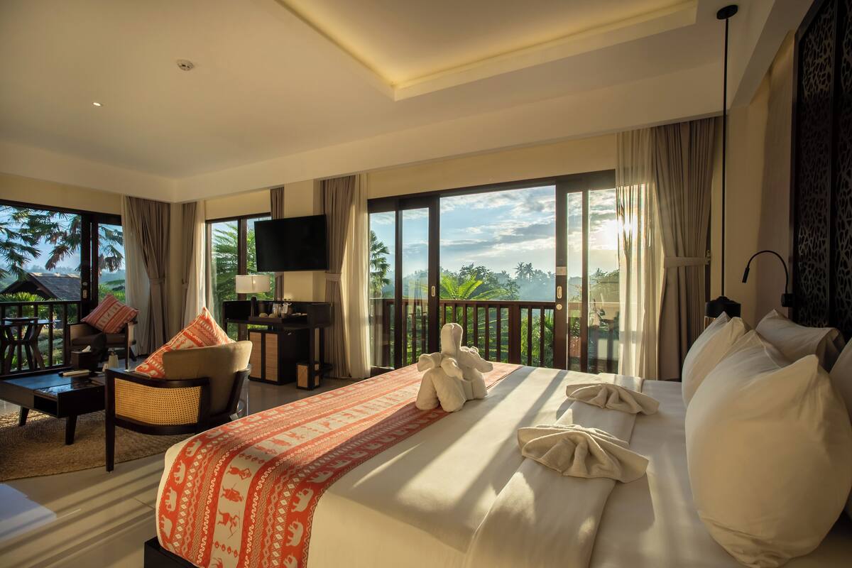 Quarto de hotel com uma cama grande, uma escrivaninha, uma cadeira e uma televisão. O quarto tem janelas amplas com vista para palmeiras e o céu.