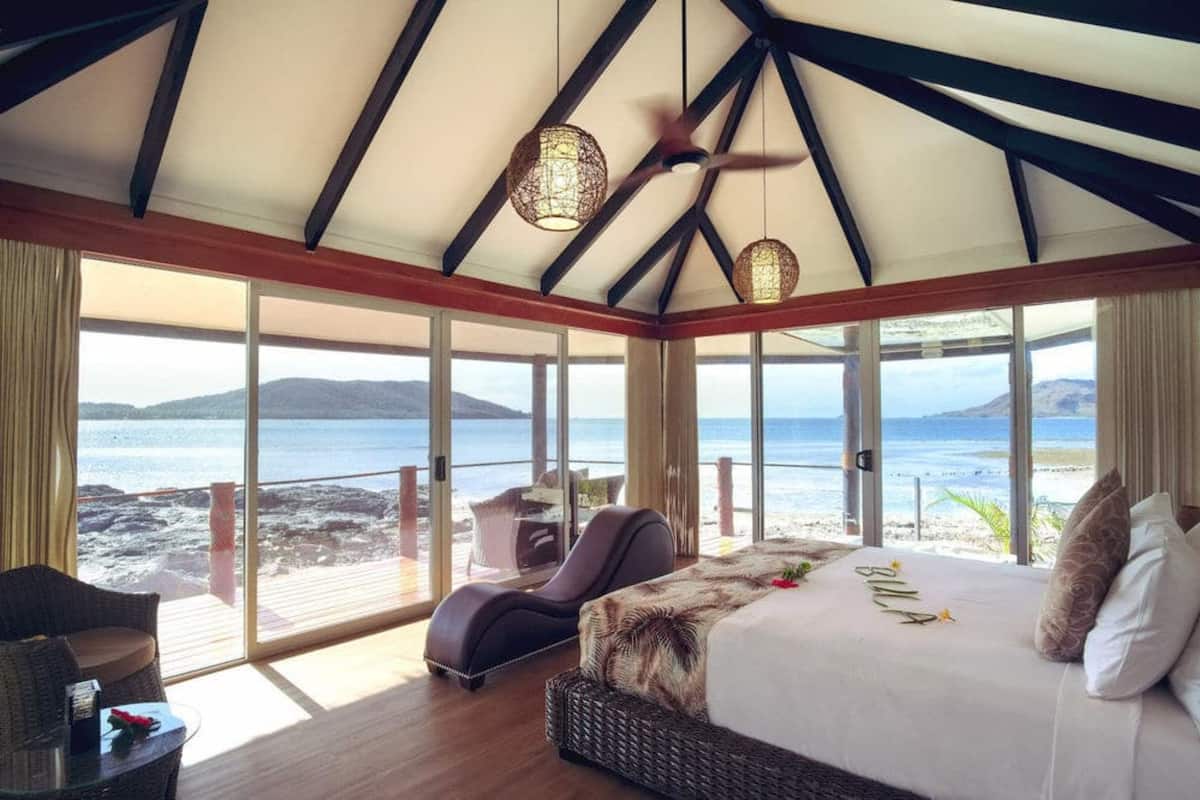 Ein Schlafzimmer mit einem großen Bett, einer Liege und Blick auf Strand und Meer.