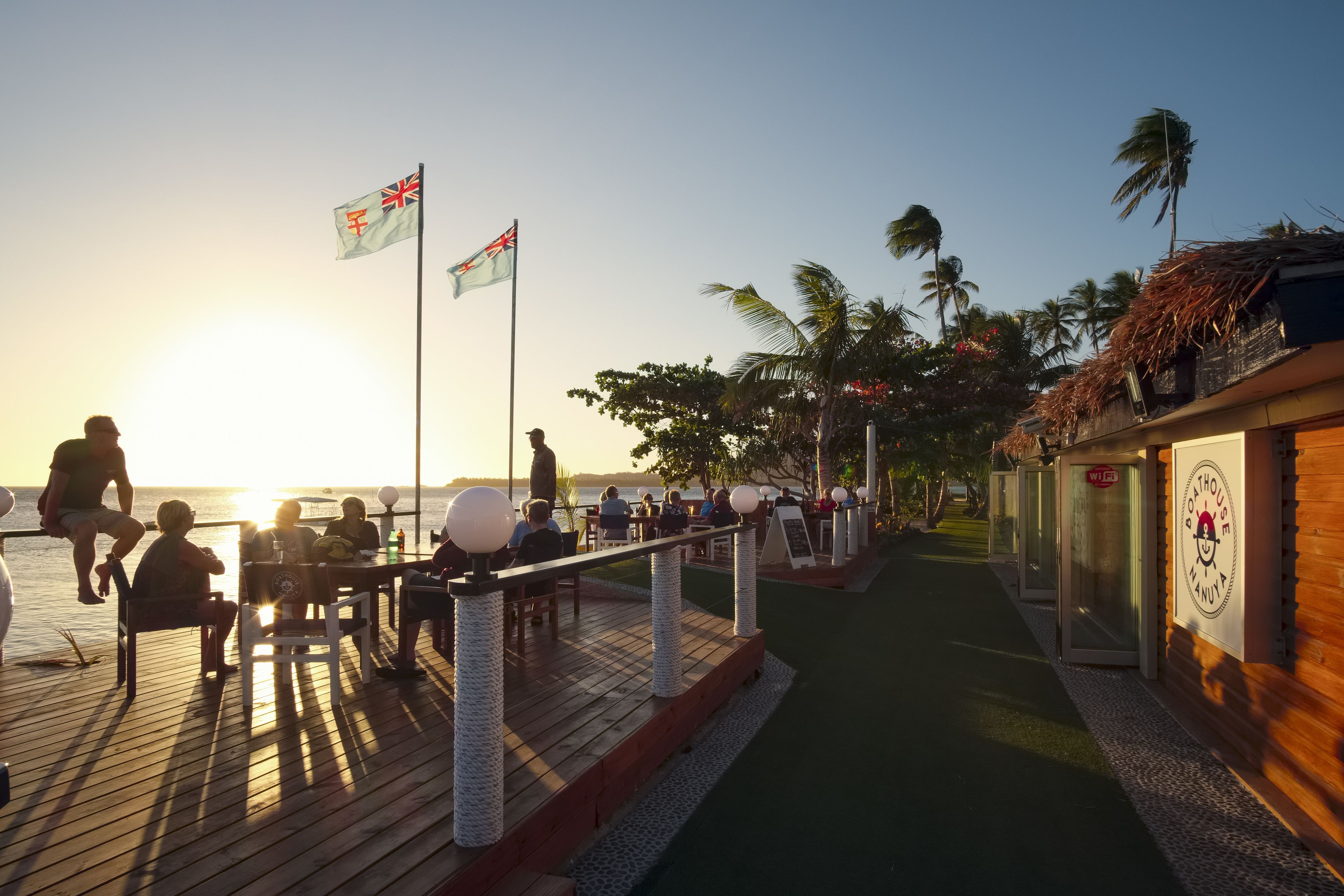 A wooden deck with tables and chairs, overlooking a body of water, with flags and palm trees in the background.