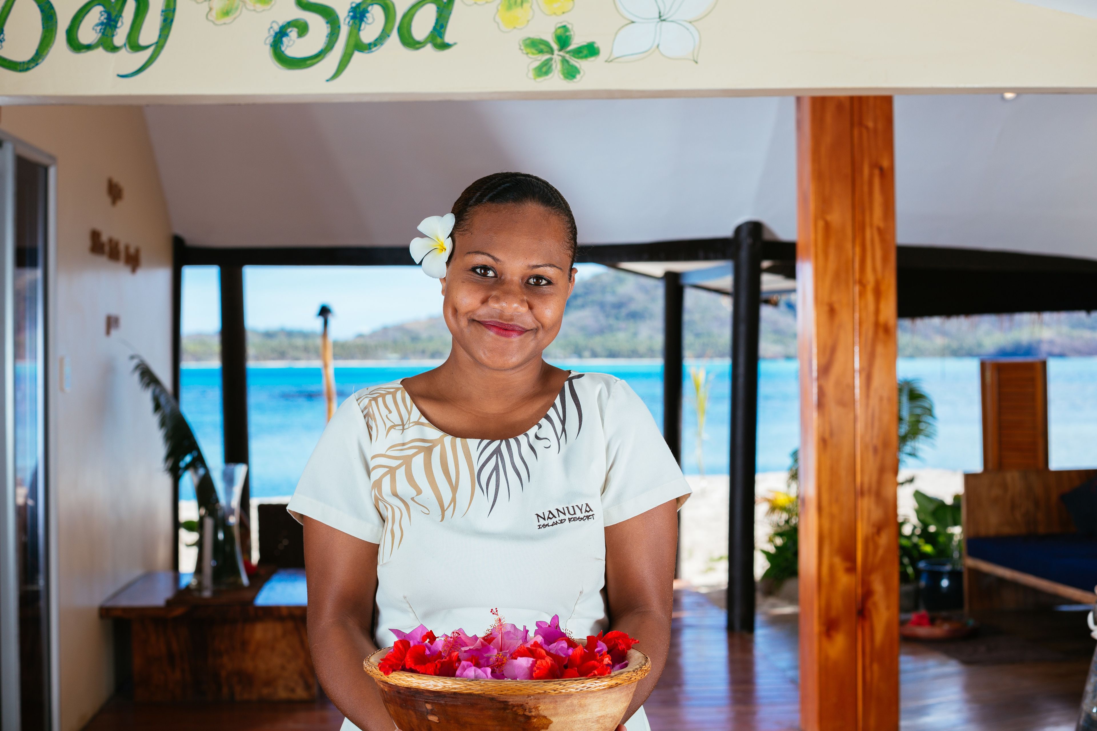 A person holding a bowl of flowers in a room with a view of the sea, a wooden structure, and a sign that reads "Day Spa".