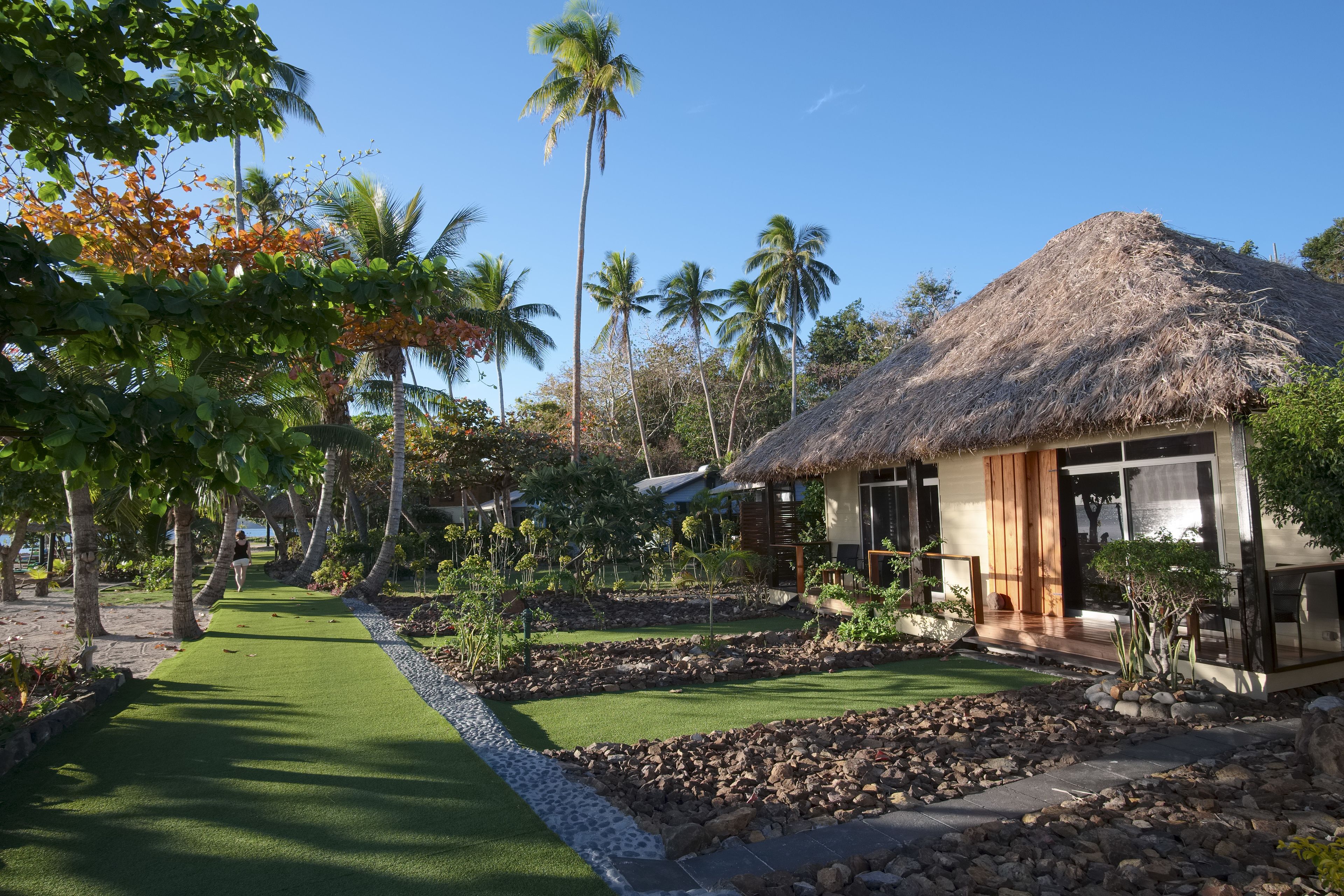 A thatched-roof house with a wooden deck, surrounded by tropical vegetation and palm trees.
