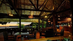 Un restaurant en plein air avec un toit de chaume, des tables et des chaises en bois, offrant une vue sur une étendue d’eau.