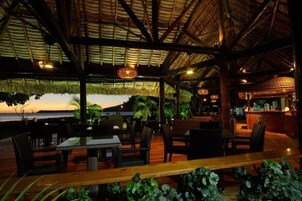 Un restaurante al aire libre con techo de paja, mesas y sillas de madera, con vistas a un cuerpo de agua.