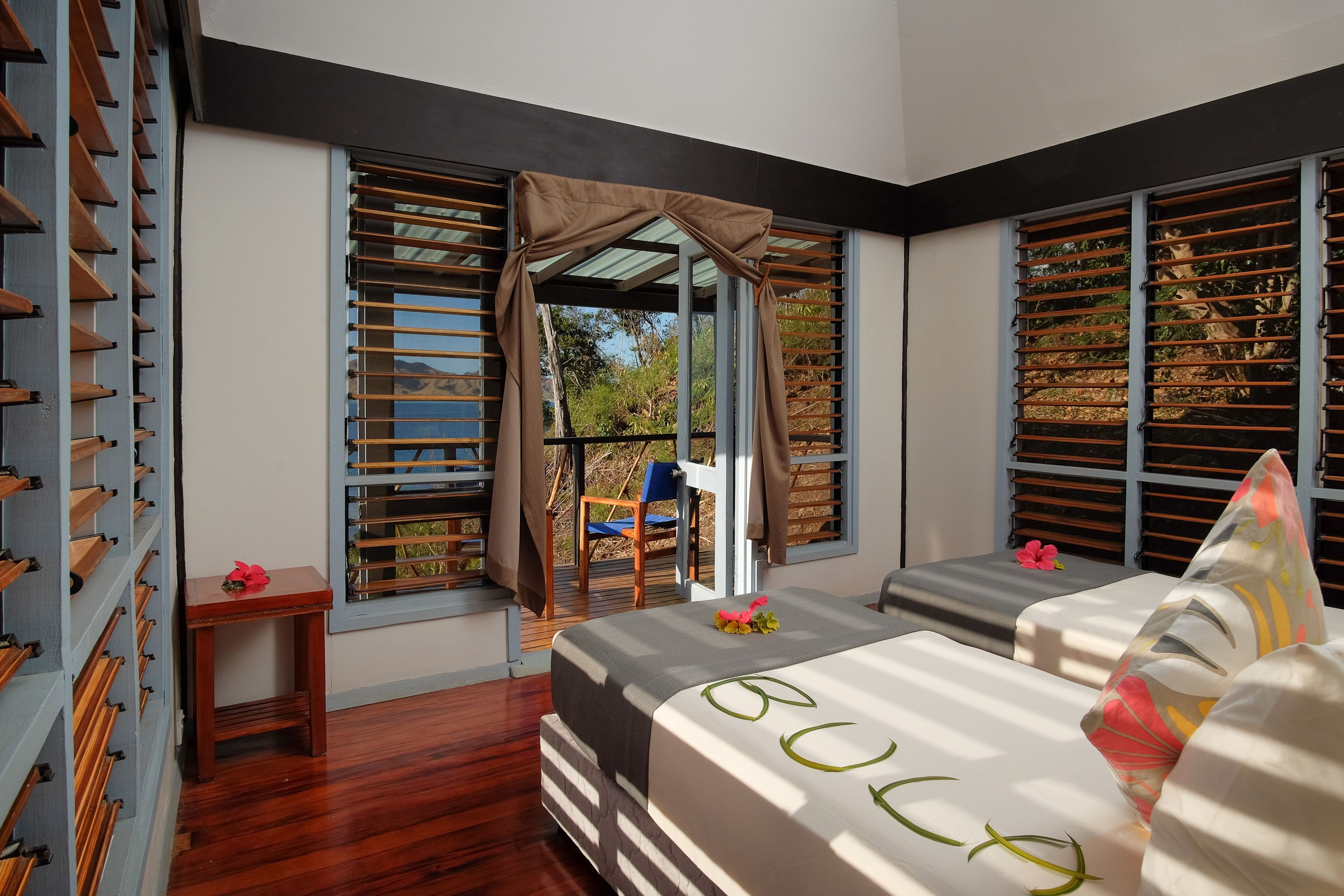 Quarto de hotel moderno com uma cama grande, uma mesa pequena e vista para o exterior através de uma janela com persianas de madeira.