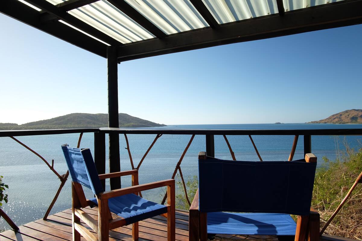 Eine Holzdar Terrasse mit zwei blauen, gepolsterten Stühlen, die auf ein ruhiges Meer und ferne Hügeln blicken.