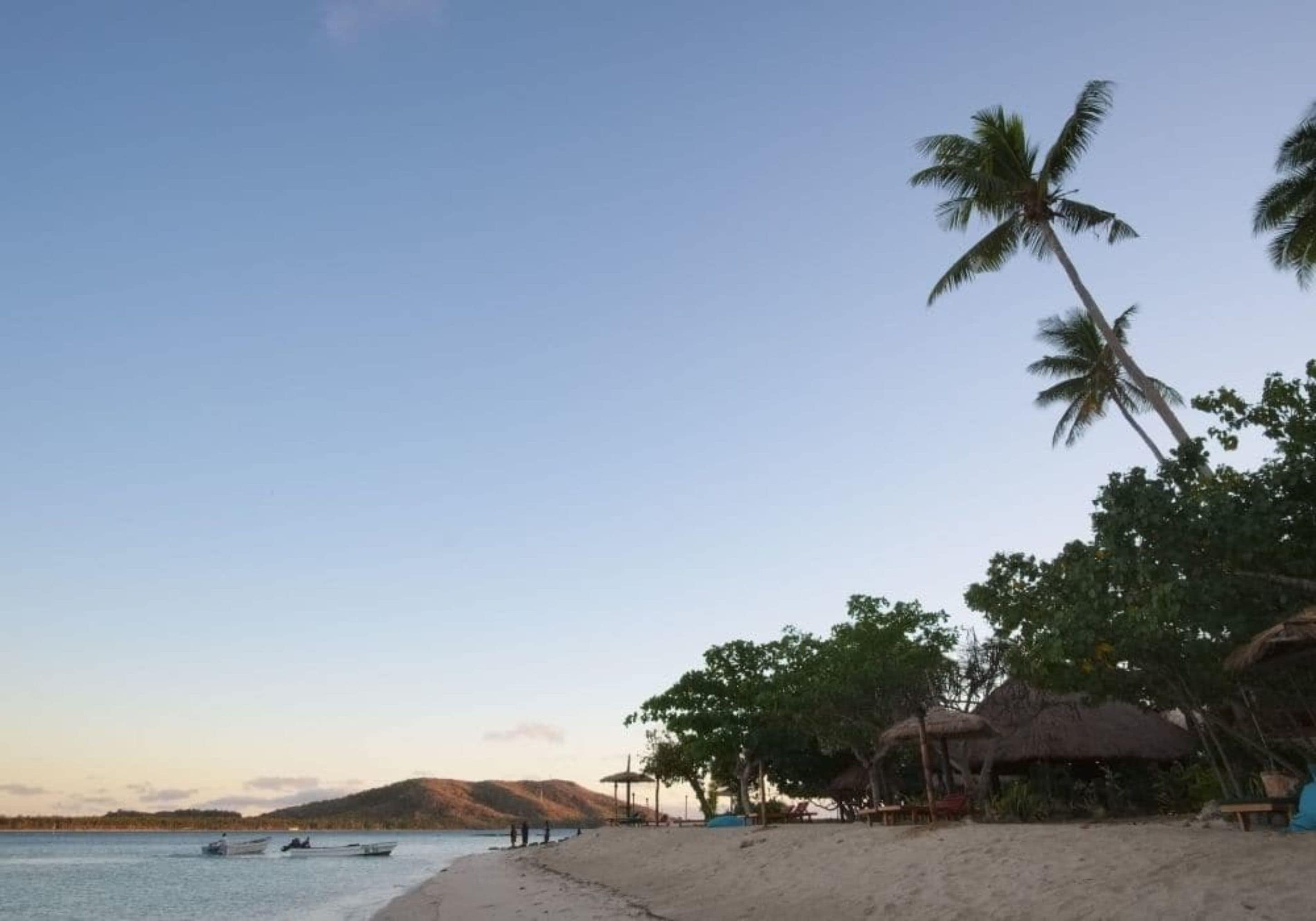 A beach with thatched-roof huts, palm trees, and boats on calm water.