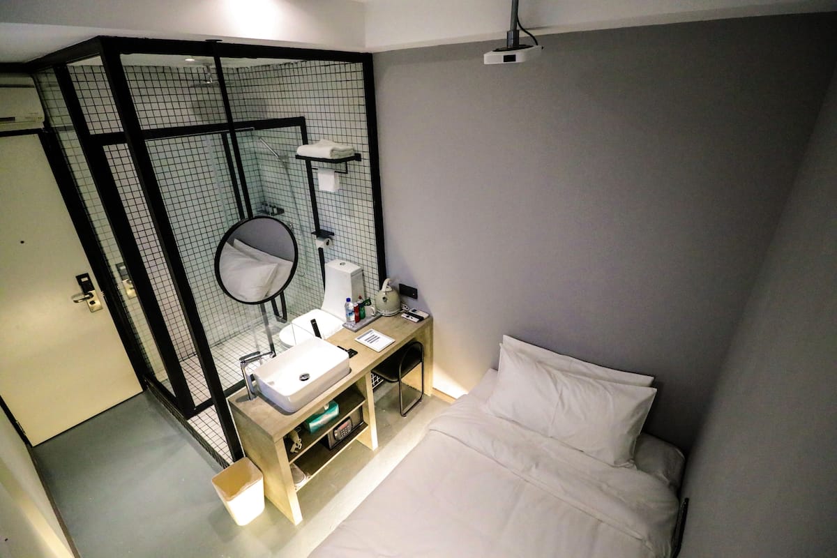 Habitación de hotel con una cama, una mesita de noche con lámpara, un espejo y zona de ducha con perchero para toallas.