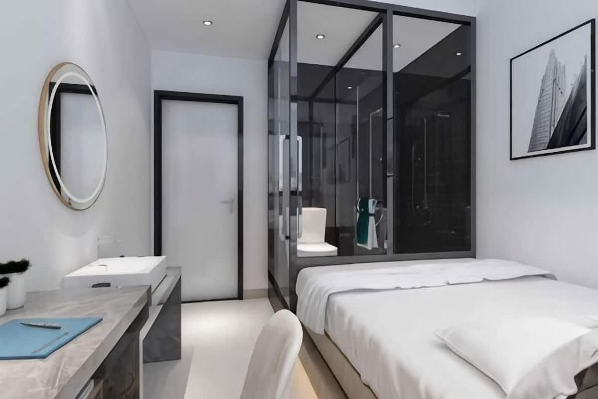 Una habitación de hotel moderna con ducha envuelta en vidrio, tocador de mármol, cama con ropa blanca y un cuadro en la pared.