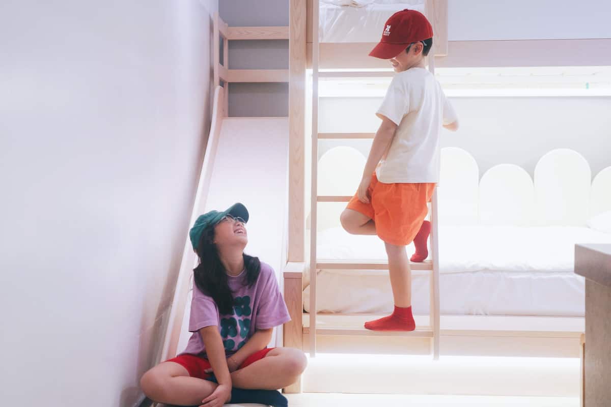 Una cama litera con escalera, un niño de pie en la cama superior y otro niño sentado en el suelo.