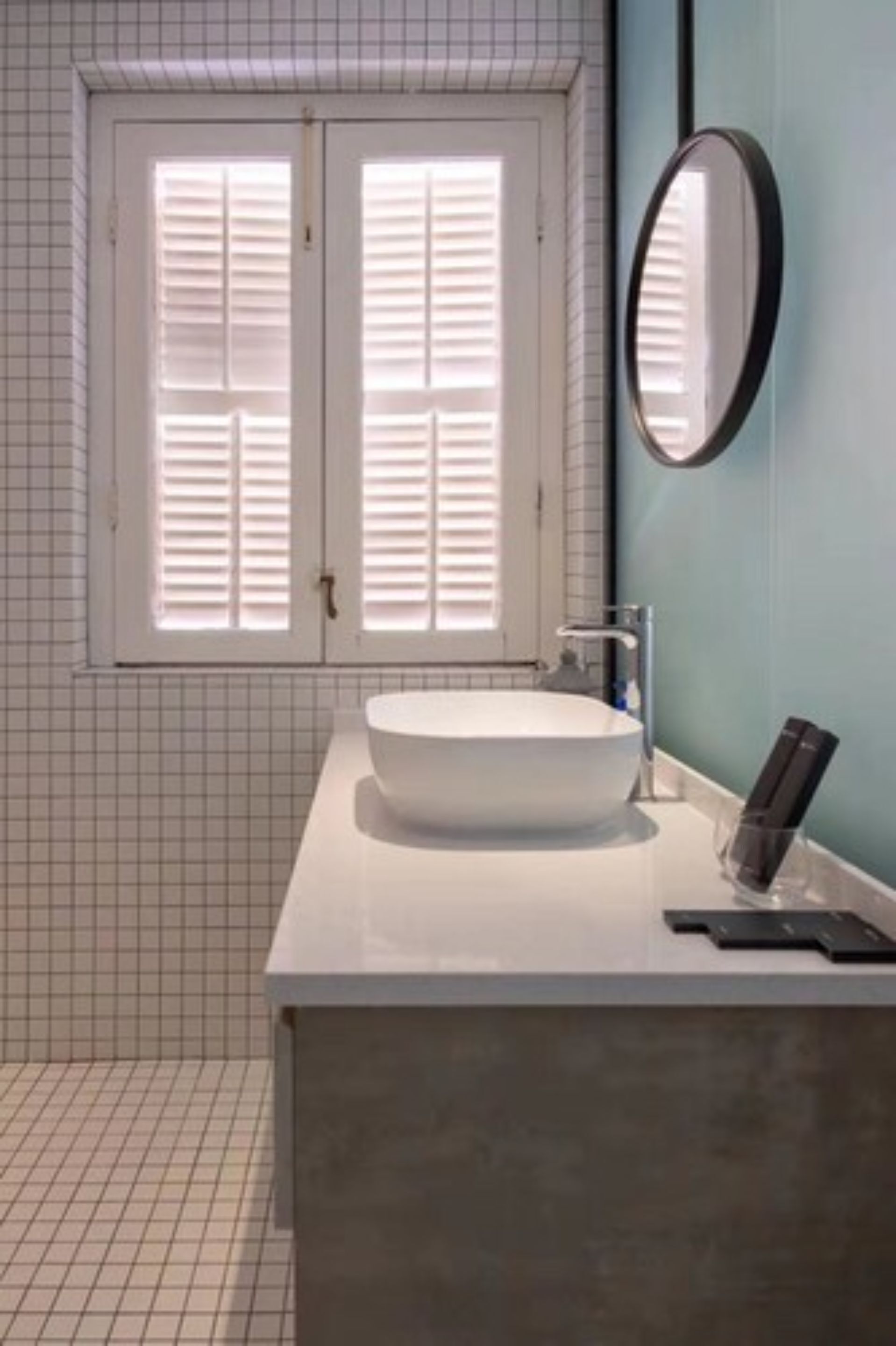 Casa de banho com lavatório branco, espelho e janela com persianas.