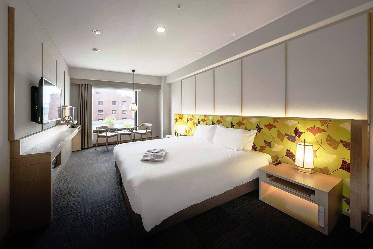 A hotel room with a bed, bedside table, lamp, TV, and a view of the city.