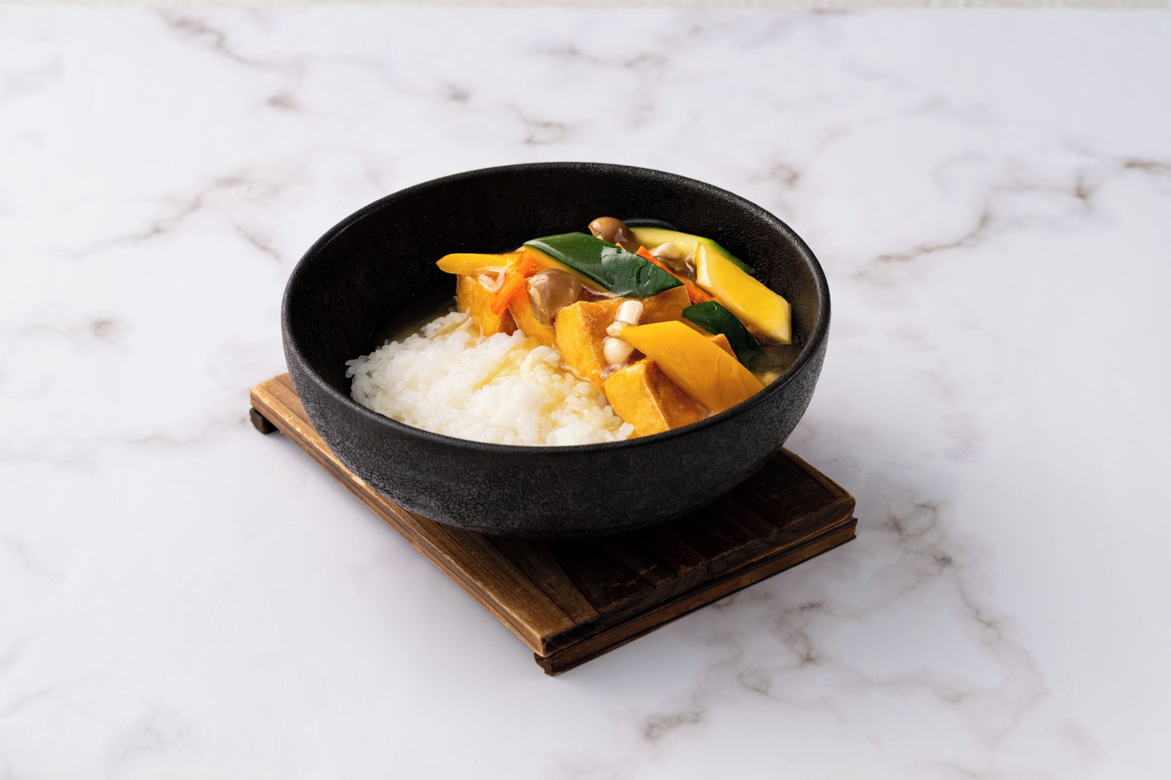 A bowl of white rice with vegetables and tofu on a dark bowl placed on a wooden coaster.