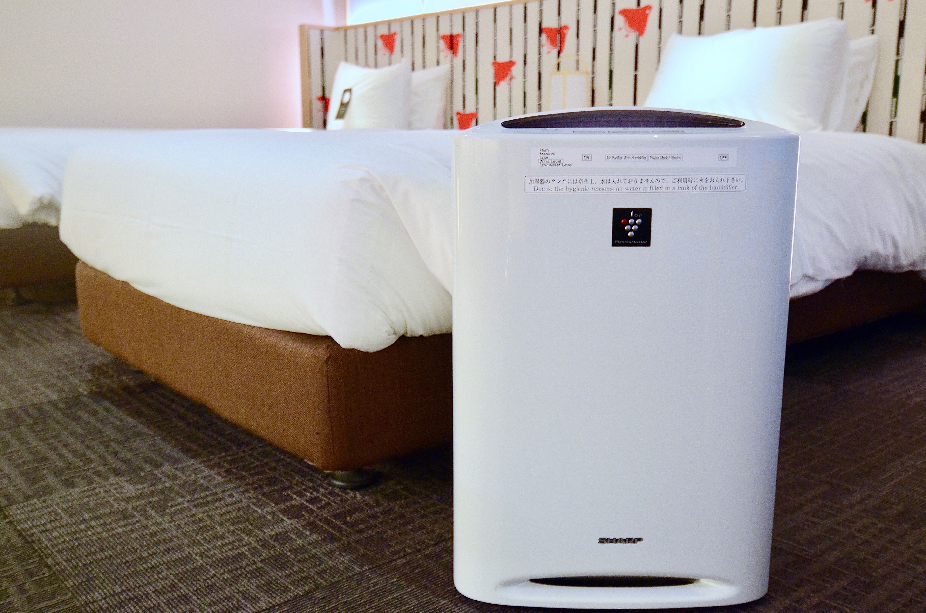 A white humidifier on a carpeted floor with a bed in the background.