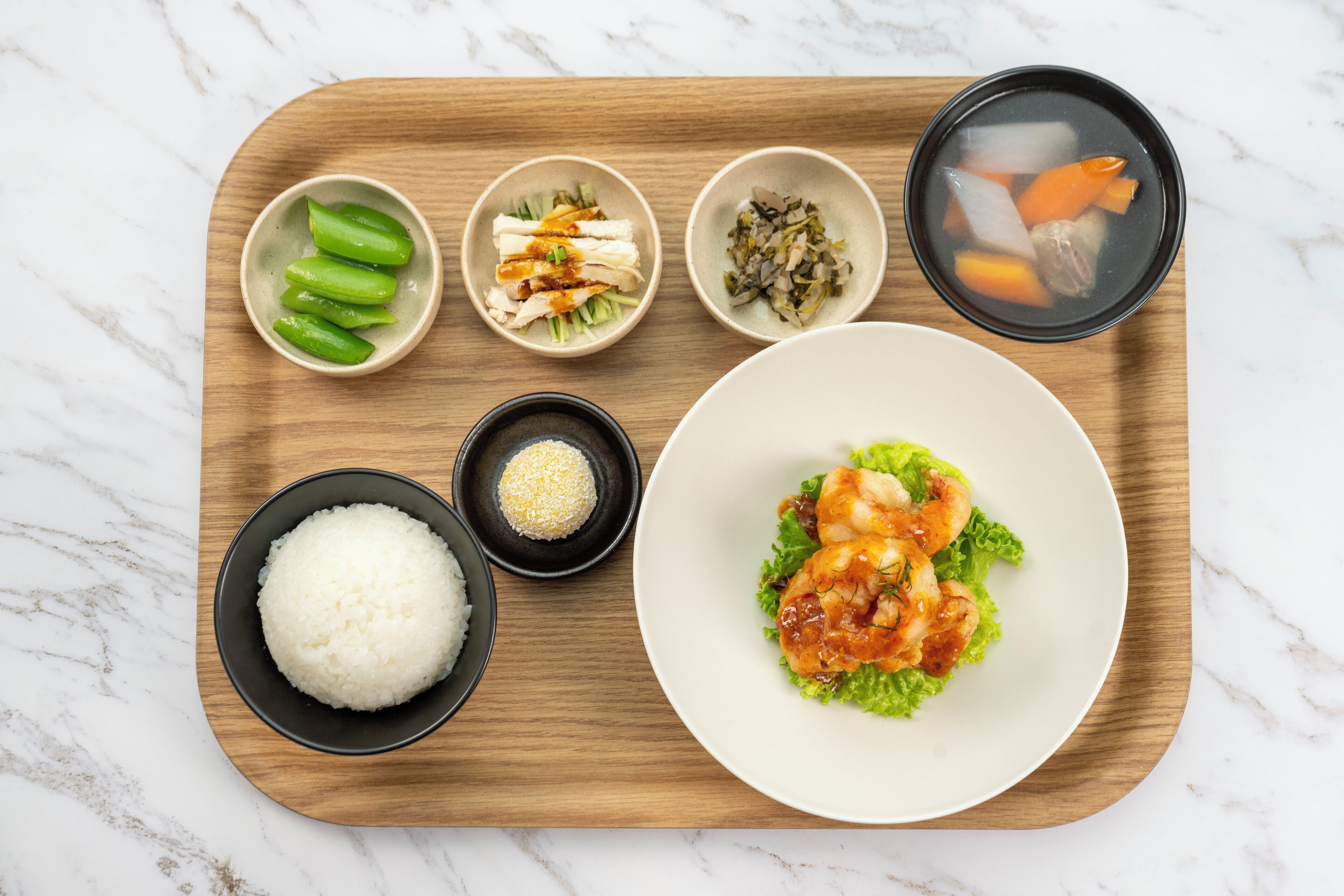 Una bandeja de madera con diversos alimentos, incluyendo arroz, verduras verdes y un tazón de sopa.