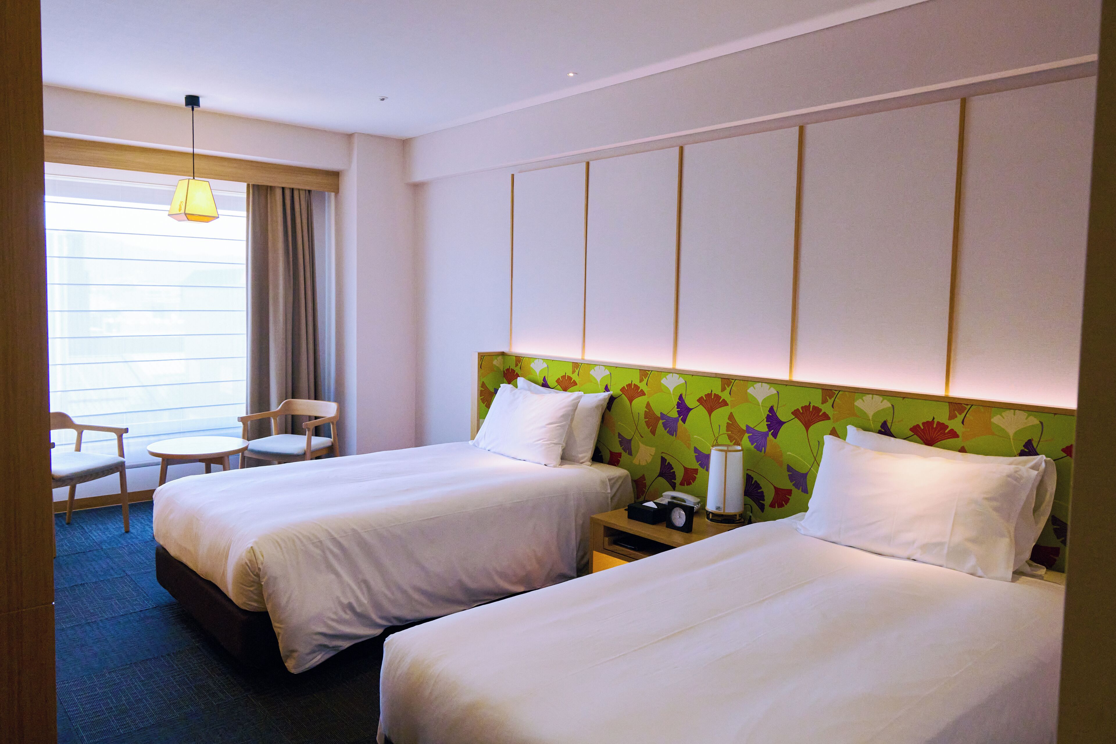 Quarto de hotel com duas camas, uma mesa pequena, cadeiras, uma luminária e uma janela com cortinas.