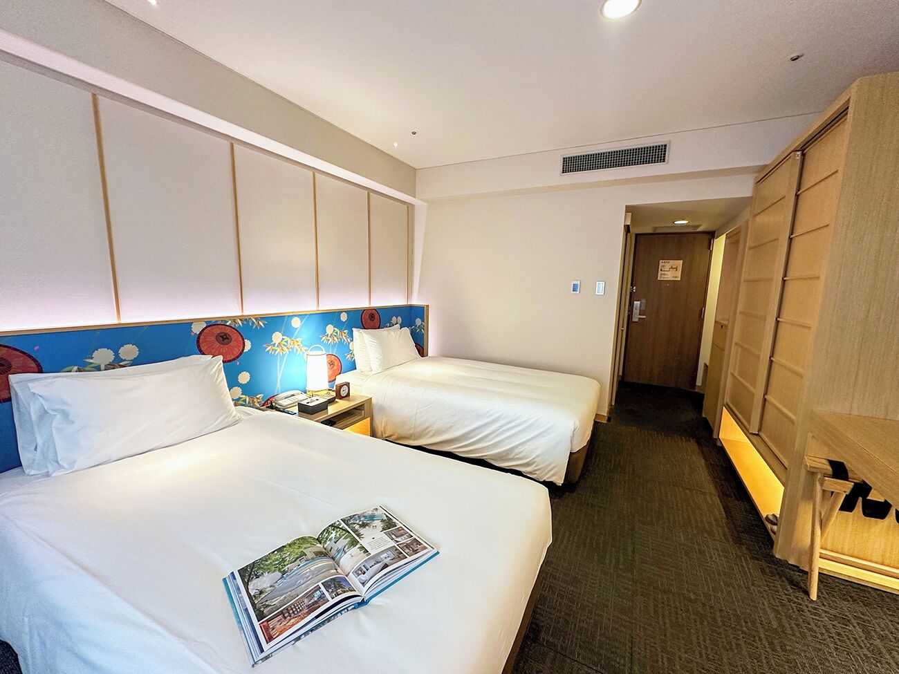 A hotel room with two beds, a nightstand with a lamp, a wardrobe, and a door leading to another room.