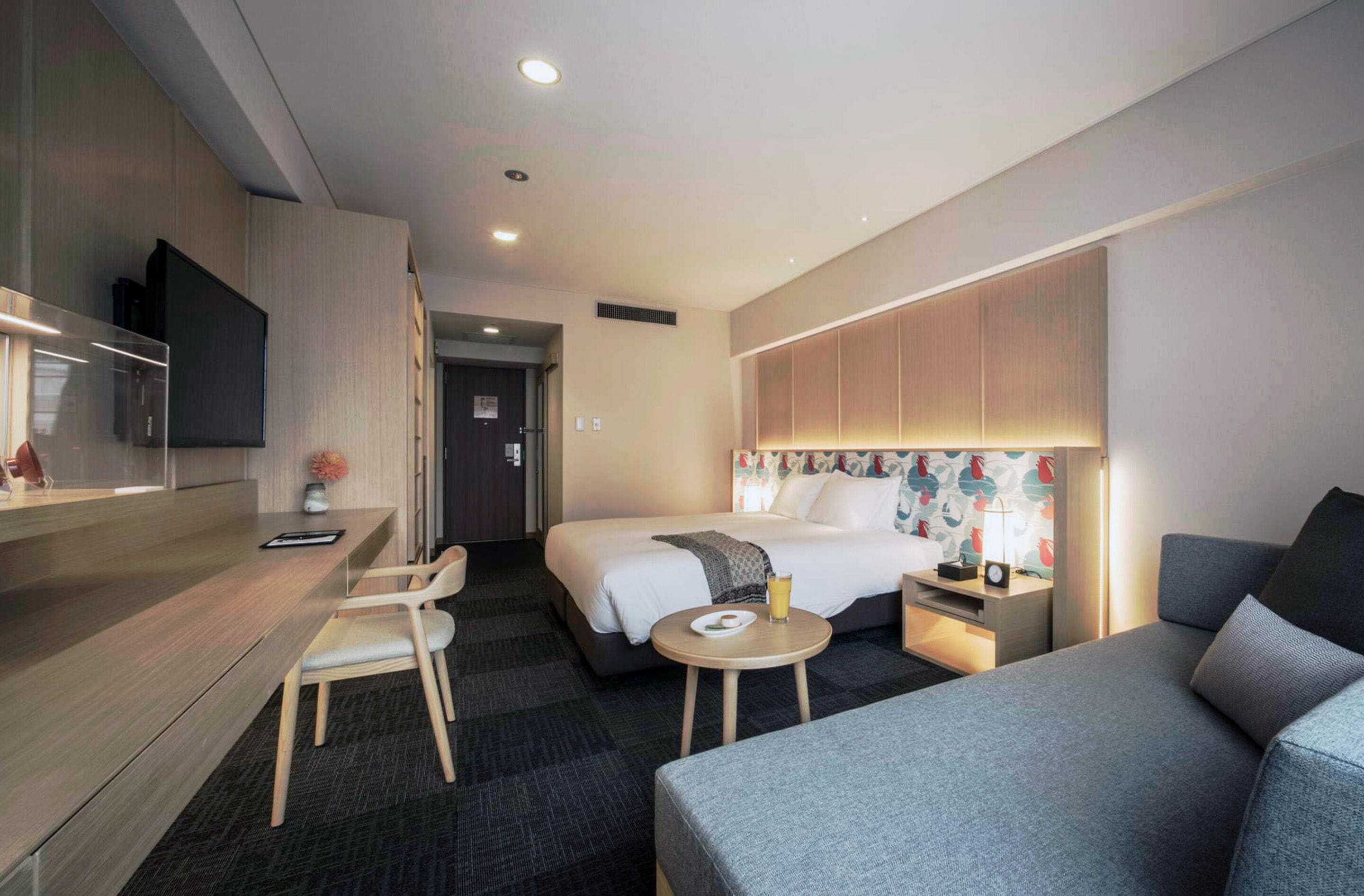 Una habitación de hotel moderna con una cama, un escritorio con una silla, un sofá y un televisor montado en la pared.