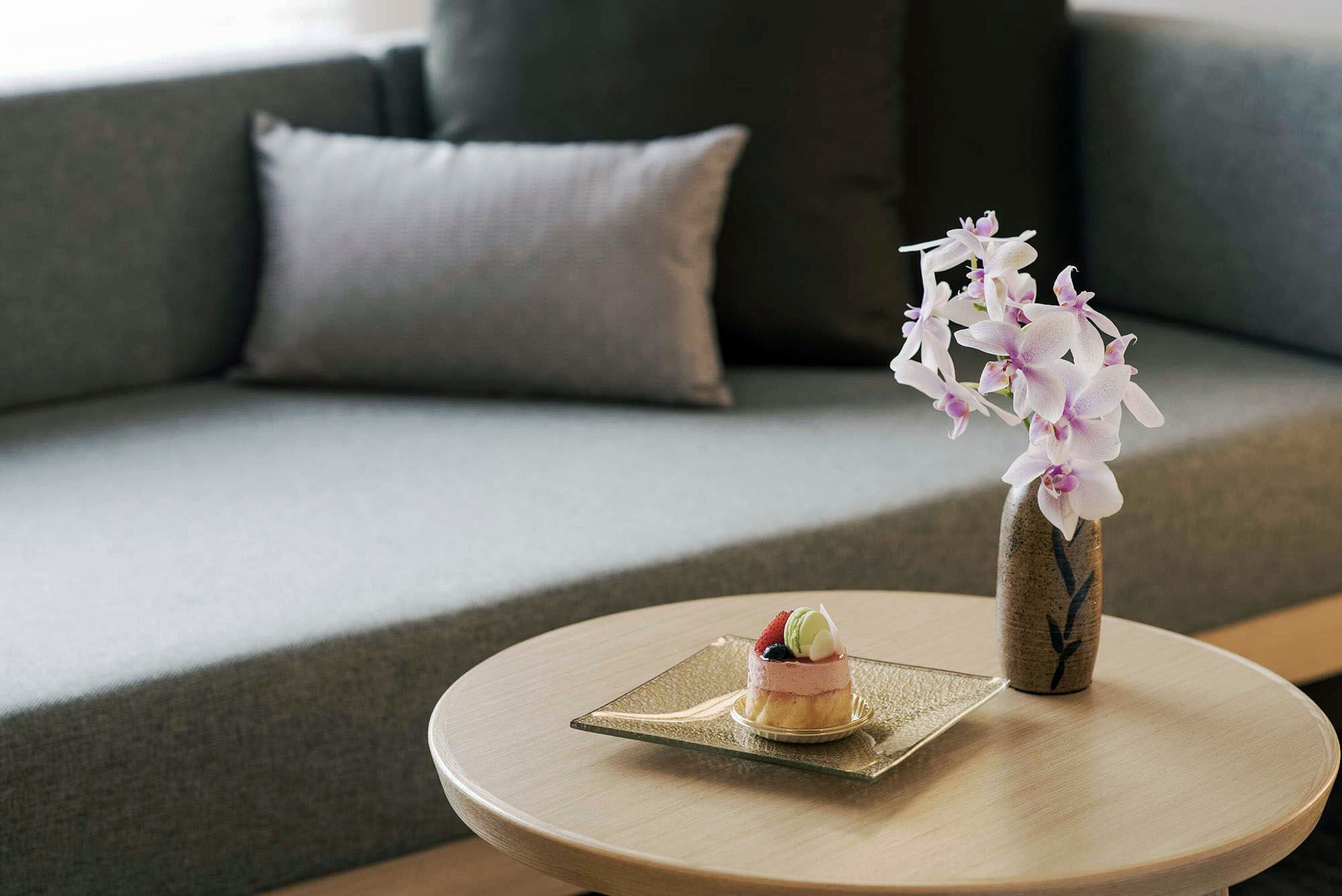 A dessert on a glass plate on a round table, a vase with flowers, and a grey sofa with cushions.