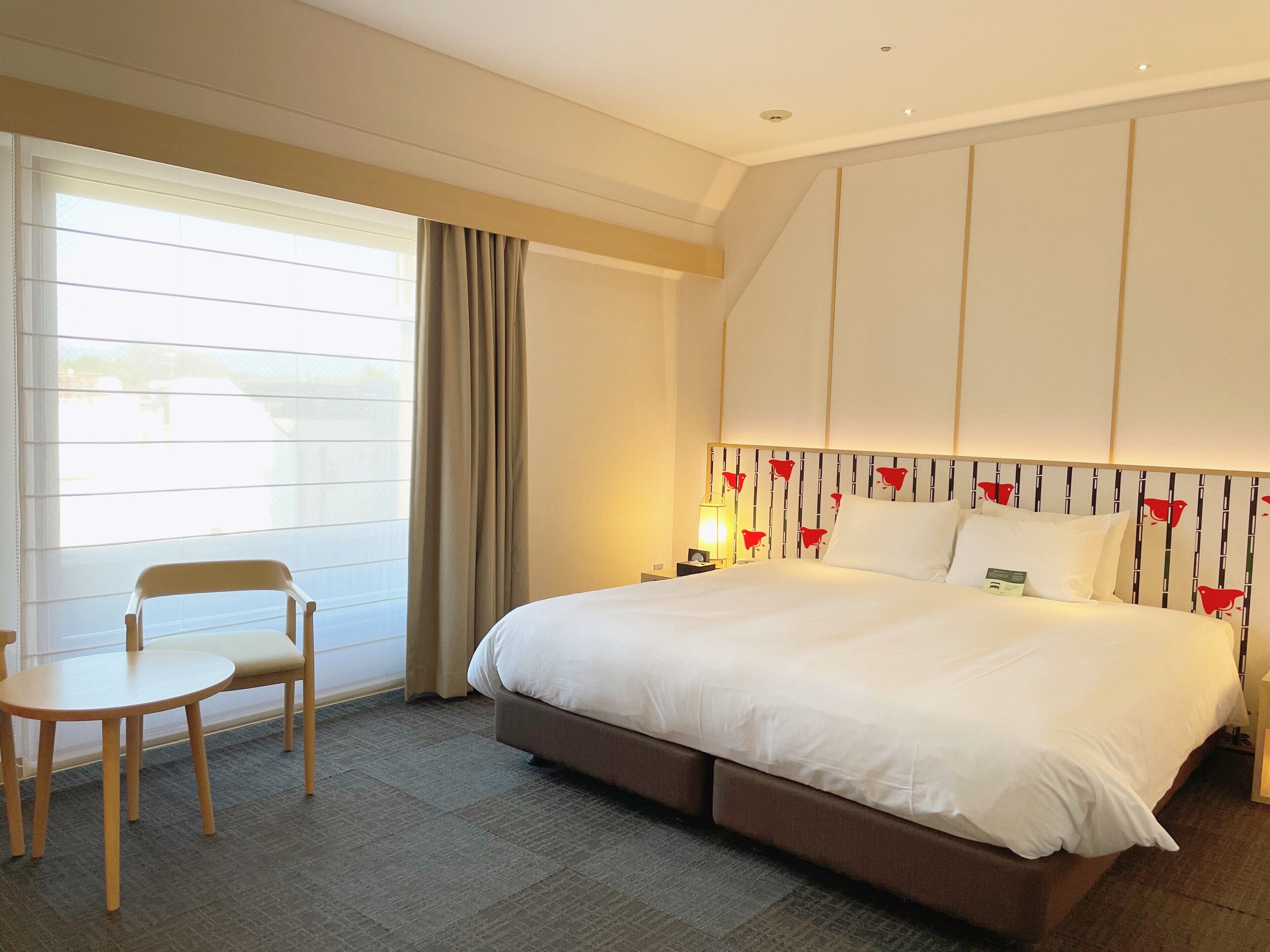 A hotel room with a large bed, a bedside table, a chair, and a window with curtains.