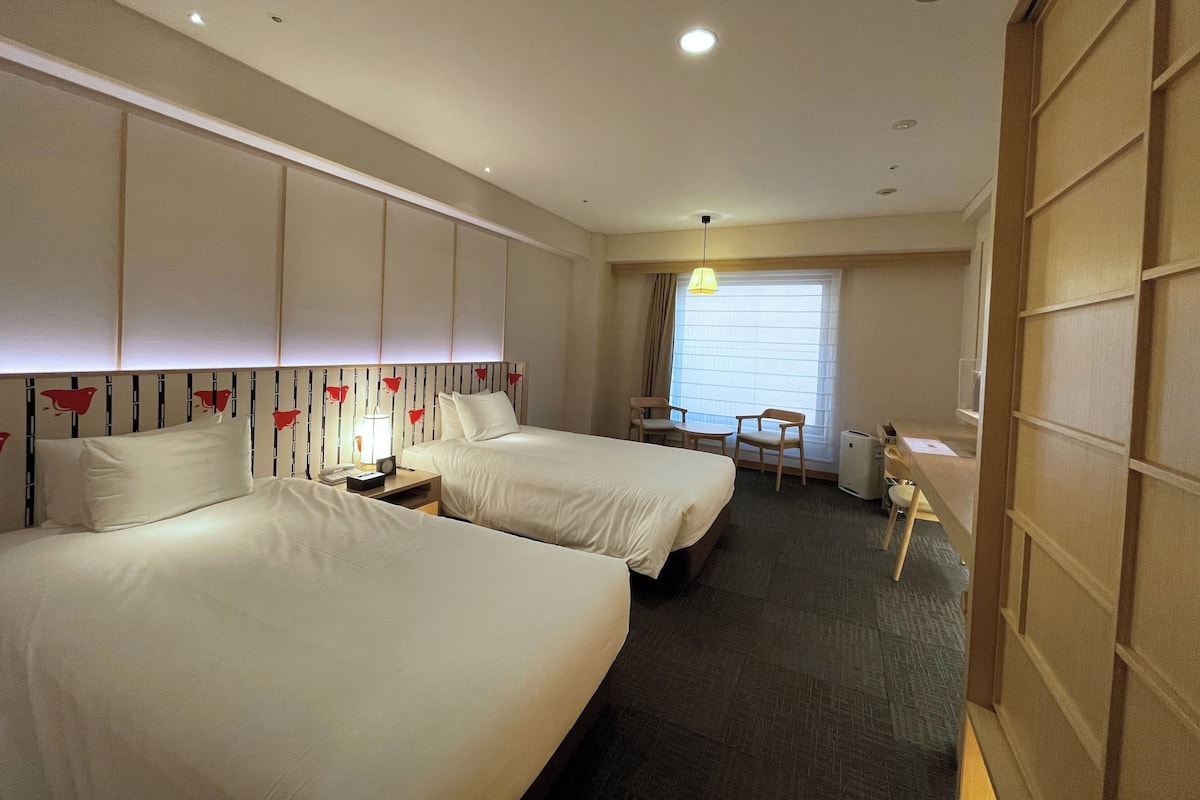 A hotel room with two beds, a desk, and a chair. There is a window with blinds, a lamp, and a ceiling light.