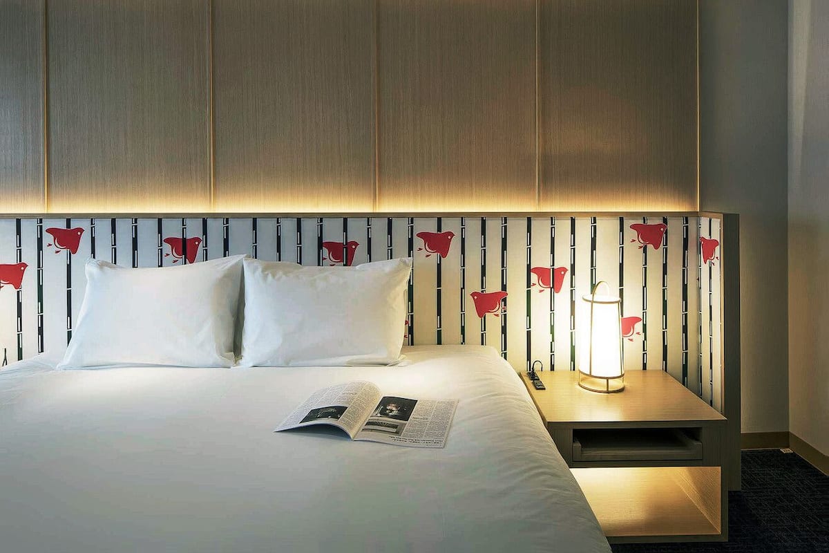 A hotel room with a bed, pillows, a bedside table with a lamp, and a wall with vertical panels.