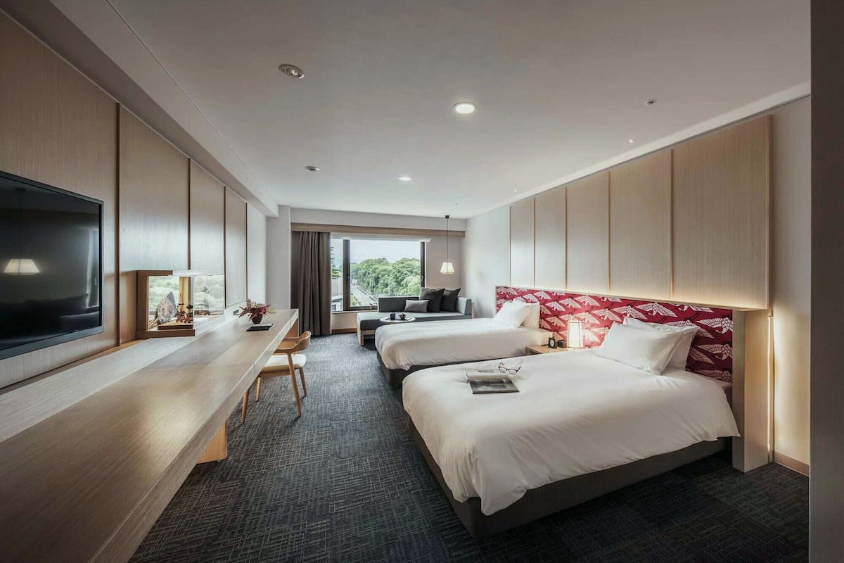 A hotel room with two beds, a desk, a TV, and a view of the outdoors.