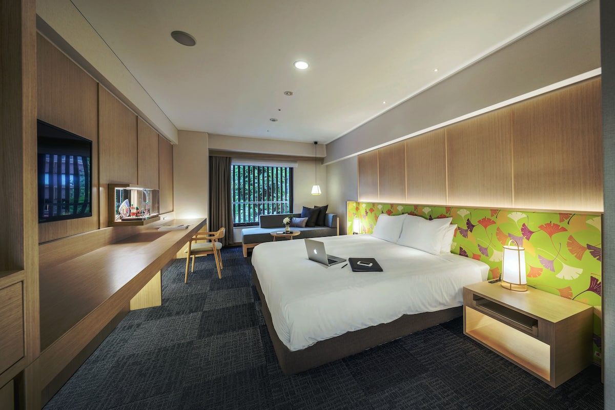 A hotel room with a bed, a desk, a chair, a TV, and a window with a view of trees.
