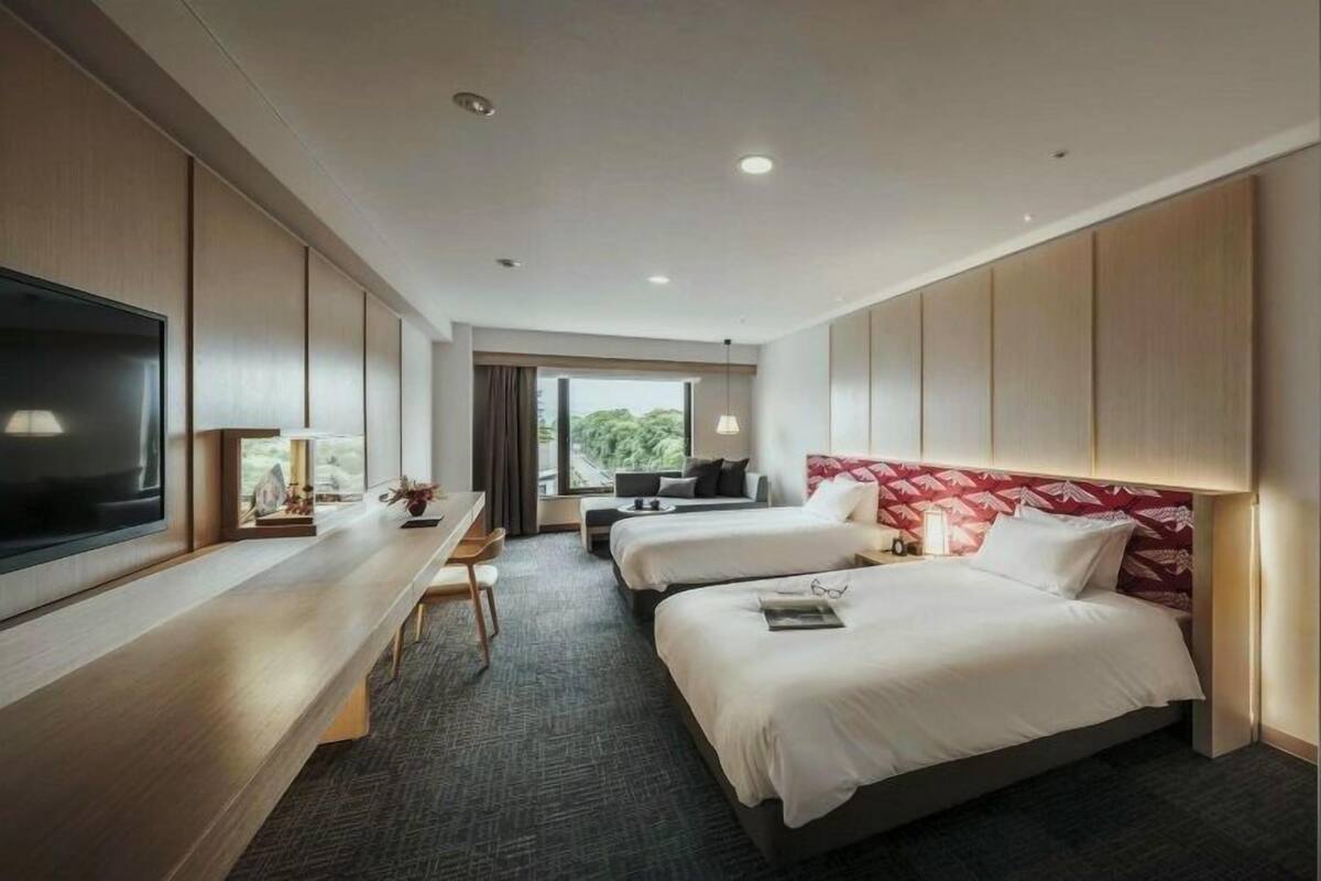 A hotel room with two beds, a TV, a desk, and a window with a view.