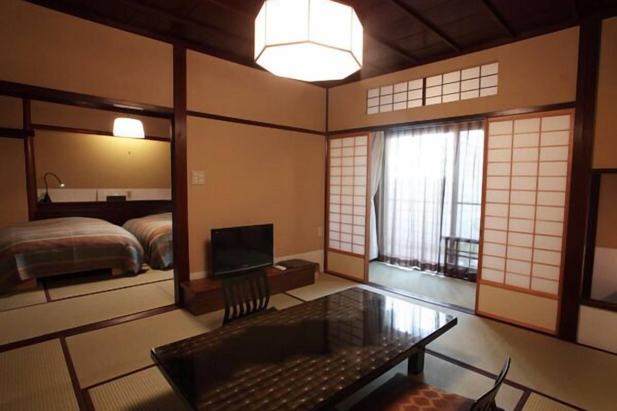 Une pièce de style japonais traditionnel, avec un sol recouvert de tatamis, une petite table basse avec un coussin, un téléviseur et des portes coulissantes donnant sur l’extérieur.
