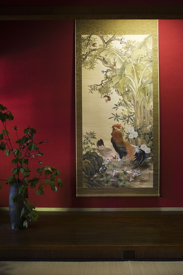 A framed traditional painting of a rooster on a red wall.