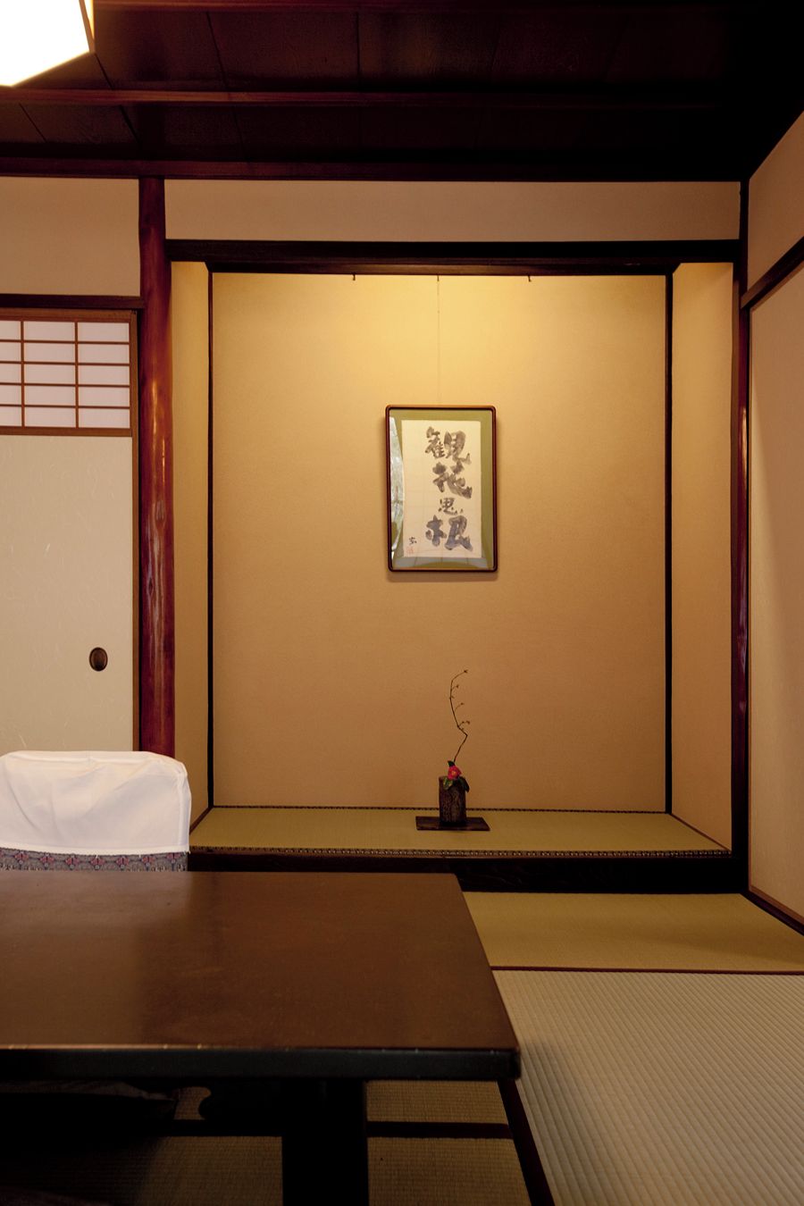 Um quarto japonês tradicional com tatames, uma mesa baixa, uma obra de caligrafia emoldurada e um pequeno vaso com um galho.