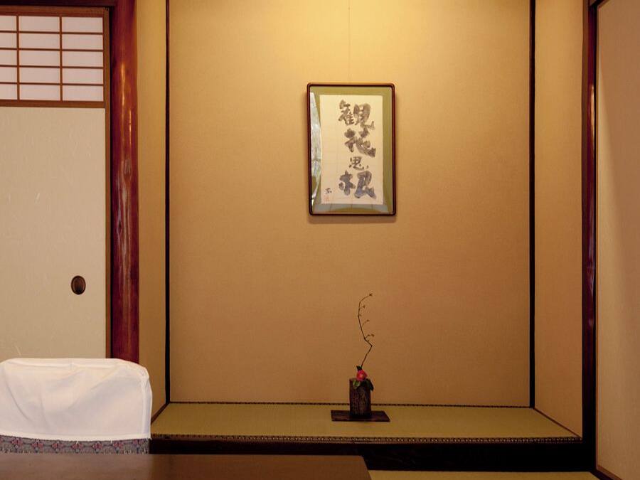 Um quarto japonês tradicional com tatames, uma mesa baixa, uma obra de caligrafia emoldurada e um pequeno vaso com um galho.