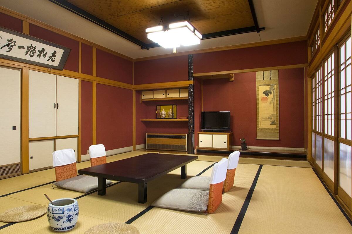 Une pièce de style japonais traditionnel, avec un sol recouvert de tatamis, une table basse, des coussins, un téléviseur et une porte coulissante.