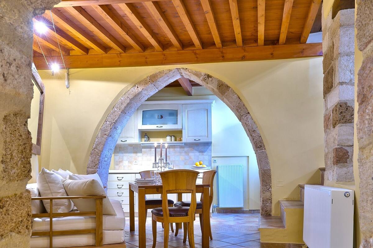 A stone archway leading to a cozy dining area with a wooden table and chairs, a sofa, and a kitchen in the background.