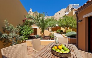 A wooden table with wicker chairs and a fruit bowl in an outdoor courtyard with potted plants and a view of buildings.